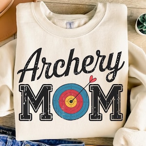 Puede incluir: Una sudadera de color crema con el texto "Archery Mom" en negro con un aspecto desgastado. El texto está centrado en la sudadera e incluye un corazón rojo y un blanco con anillos rojos, amarillos, azules y blancos.