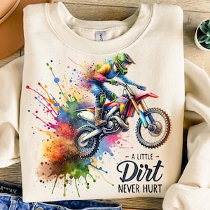 Puede incluir: Sudadera color crema con una ilustración de acuarela de un piloto de motocross en una moto roja y azul. El texto "A Little Dirt Never Hurt" está debajo de la imagen. El diseño incluye vibrantes salpicaduras de pintura.