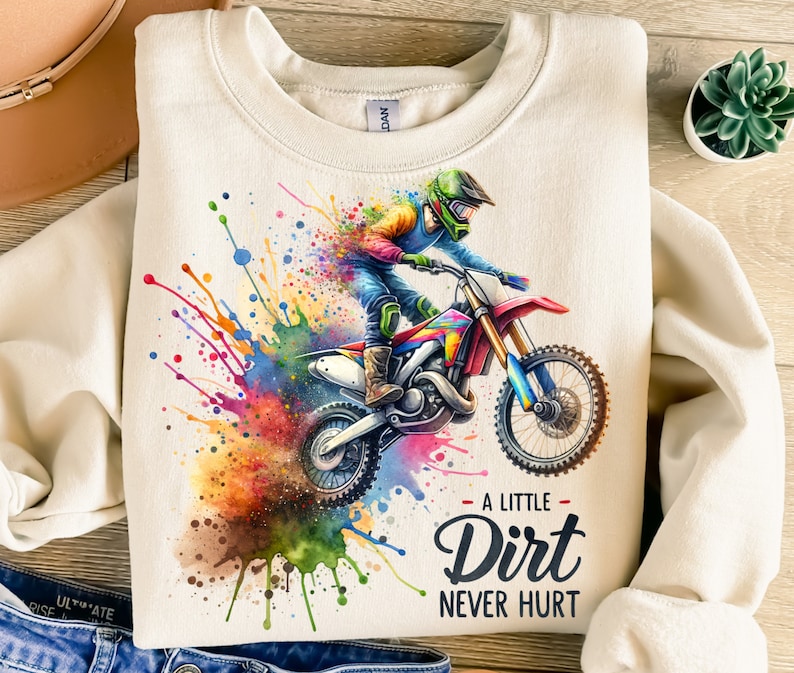 May include: Cream-colored sweatshirt featuring a colorful watercolor illustration of a motocross rider on a red and blue dirt bike. The text "A Little Dirt Never Hurt" is below the image. The design includes vibrant paint splatters.