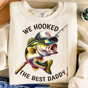 May include: A beige sweatshirt with the text "We hooked the best daddy" in brown lettering. A cartoon bass fish wearing a baseball cap with an American flag is holding a fishing rod.