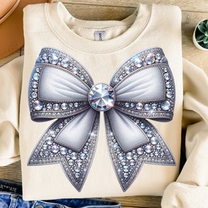 May include: A cream colored sweatshirt with a large white bow adorned with sparkling silver rhinestones. The bow has a large, clear rhinestone in the center.