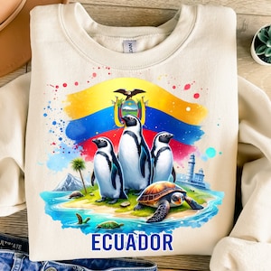 Op de afbeelding: Een beige sweatshirt met een kleurrijk aquarelontwerp met de vlag van Ecuador, drie pinguïns, een zeeschildpad en een vuurtoren. De tekst "ECUADOR" is onder het ontwerp gedrukt.