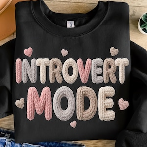 May include: Black sweatshirt with the words "INTROVERT MODE" in a textured, knitted style. The letters are in shades of pink, gray, and beige, with small pink hearts scattered around the text. A casual, comfortable design.
