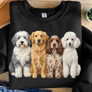 May include: Black sweatshirt featuring a graphic of four different dog breeds: a gray and white Old English Sheepdog, a golden retriever, a brown and white Cocker Spaniel, and a white poodle. The dogs are sitting side by side.