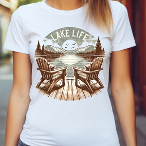 May include: White t-shirt featuring a lakeside scene with two wooden Adirondack chairs on a dock, mountains, trees, and the text "LAKE LIFE". The design is in shades of brown, green, and blue.
