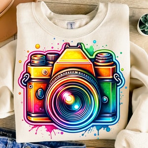 以下が含まれることがあります： クリーム色のスウェットシャツに、ビンテージカメラのカラフルなグラフィックがプリントされています。カメラはネオンカラーで縁取られ、虹色のレンズが付いています。背景は、ピンク、ブルー、グリーン、イエローの水彩画です。