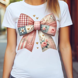 May include: White t-shirt featuring a large, decorative bow graphic. The bow is a patchwork design with pink, blue, and red patterns, including gingham, floral, and striped elements. Gold heart accents are scattered around the bow.