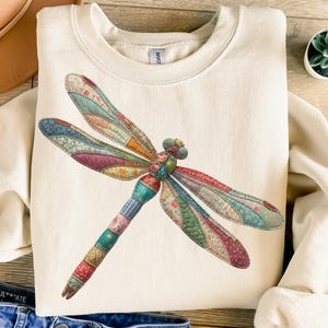 May include: Cream-colored sweatshirt featuring a large, colorful dragonfly design. The dragonfly has patchwork-style wings in shades of pink, blue, green, and orange. The body is a mix of colors. The sweatshirt has a crew neck and long sleeves.