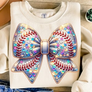 Puede incluir: Una sudadera blanca con un gran lazo brillante iridiscente. El lazo está hecho de lentejuelas y tiene un diseño de béisbol.