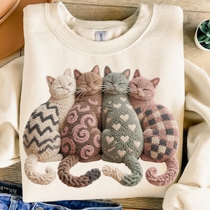 Könnte beinhalten: Cremefarbenes Sweatshirt mit einer Grafik von vier stilisierten Katzen. Jede Katze hat ein einzigartiges Muster: Zickzack, Wirbel, Herzen und Quadrate. Die Katzen sind in Grau-, Braun- und Rosatönen gehalten, mit geschlossenen Augen. Das Sweatshirt hat einen Rundhalsausschnitt.