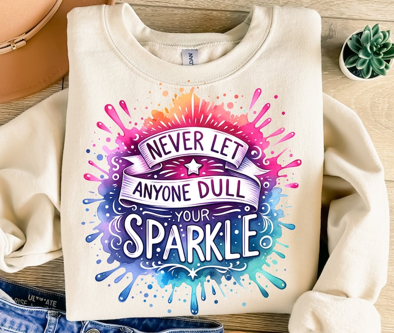 May include: A cream coloured sweatshirt with a watercolour design of colourful paint splatters. The design features the text "Never let anyone dull your sparkle."