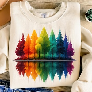 Puede incluir: Sudadera color crema con un vibrante diseño de bosque arcoíris. El gráfico muestra árboles en tonos rojos, naranjas, amarillos, verdes, azules y morados, reflejados en el agua. El diseño está centrado en la parte delantera de la sudadera.