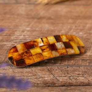 Puede incluir: Una pinza para el pelo de ámbar marrón y amarillo con un diseño de mosaico. La pinza está hecha de muchas piezas pequeñas de ámbar que están engastadas en un marco de metal.
