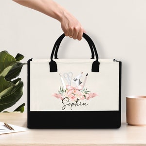 May include: A canvas tote bag with black handles and a black bottom. The bag features a floral design with scissors, a glue gun, and a pencil. The name "Sophia" is written in script below the floral arrangement.