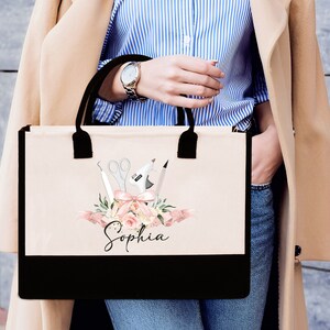 May include: A beige tote bag with black handles and a black bottom panel. The bag features a floral design with scissors, a glue gun, and other craft tools. The name "Sophia" is written in script below the design.