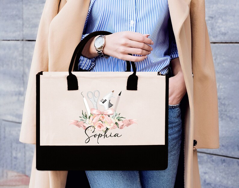 May include: A beige tote bag with black handles and a black bottom panel. The bag features a floral design with scissors, a glue gun, and other craft tools. The name "Sophia" is written in script below the design.