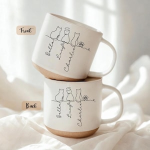May include: Two white ceramic mugs with a tan base are stacked. Each mug features a black line drawing of three cats, a flower, and the names "Bella", "Lucy", and "Charlie". The word "Front" is on one mug and "Back" on the other.