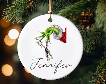 May include: White ceramic Christmas ornament with a green Grinch hand holding a pair of scissors decorated with colorful Christmas lights. The ornament has the name "Jennifer" written in cursive at the bottom.