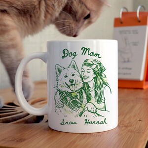 Może przedstawiać: Biały ceramiczny kubek z zieloną ilustracją kobiety i psa, z napisami "Dog Mom" i imionami "Snow Hannah". Pies ma na szyi chustę. Kubek ma zakrzywiony uchwyt.
