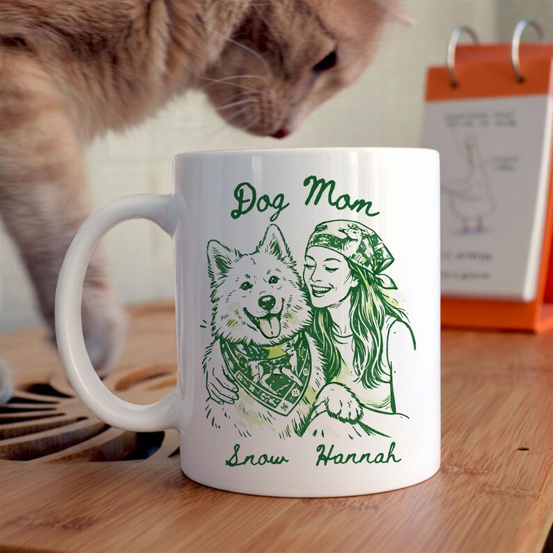 Może przedstawiać: Biały ceramiczny kubek z zieloną ilustracją kobiety i psa, z napisami "Dog Mom" i imionami "Snow Hannah". Pies ma na szyi chustę. Kubek ma zakrzywiony uchwyt.
