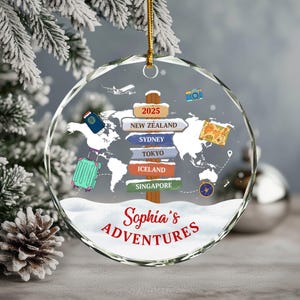 May include: A clear, round Christmas ornament with a world map design and the text "Sophia's Adventures." The ornament features travel-themed illustrations, including a suitcase, passport, and signposts pointing to various destinations.