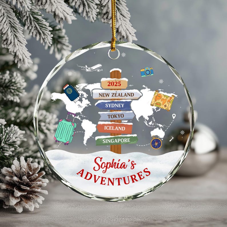 May include: A clear, round Christmas ornament with a world map design and the text "Sophia's Adventures." The ornament features travel-themed illustrations, including a suitcase, passport, and signposts pointing to various destinations.