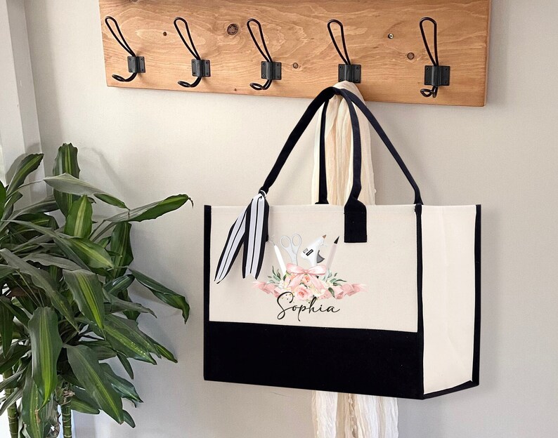 May include: A large tote bag with a black bottom and trim, and a cream-colored upper section. The bag features a floral design with scissors, a glue gun, and pencils, and the name "Sophia" in script. Black handles and a striped tassel complete the design.