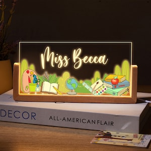 May include: A wooden light-up sign with a clear acrylic panel featuring the text "Miss Becca" and a colourful illustration of school supplies, including books, pencils, a globe, and paintbrushes. The sign is illuminated with warm white LED lights.