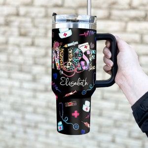 May include: A black stainless steel travel mug with a handle. The mug has a colorful design with the word "NURSE" and the name "Elizabeth". The design also includes medical symbols like a stethoscope, a syringe, and a bandage.