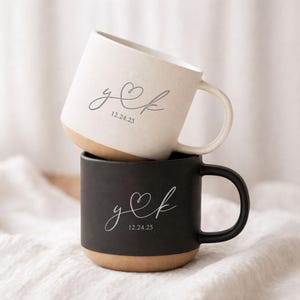 Puede incluir: Dos tazas de cerámica apiladas, una crema y otra negra. Cada taza presenta un diseño personalizado con iniciales y una fecha en letra cursiva. La base de cada taza es de color marrón natural.