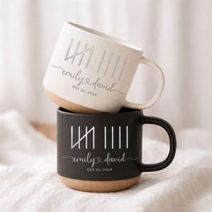 May include: Two ceramic mugs, one cream and one black, stacked together. Each mug features a unique design with vertical lines and the names "emily & david" with a heart symbol, and the date "Oct 22, 2024". The bottom of each mug is a natural tan color.