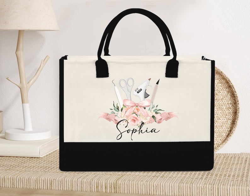 May include: A canvas tote bag with black handles and a black bottom panel. The bag features a floral design with craft tools, a pink bow, and the name "Sophia" in script.