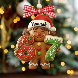 May include: A gingerbread person Christmas ornament holding the numbers 6 and 7, with the year 2025. The ornament has a red Santa hat with the name "Hannah" on it. The ornament is decorated with red, white, green, and brown colors.