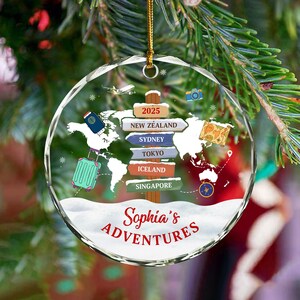 May include: A clear, round Christmas ornament with a world map design and the text "Sophia's Adventures." The ornament features a suitcase, passport, and destinations like New Zealand, Sydney, and Tokyo. A gold string is attached for hanging.