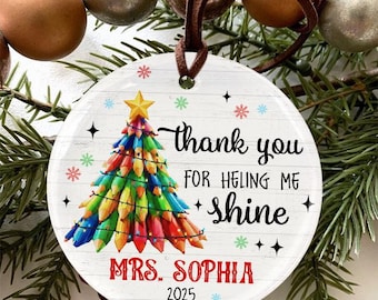 May include: A round Christmas ornament with a colorful pencil-shaped Christmas tree design. The ornament has the text "thank you for helping me shine" and "Mrs. Sophia 2025". The ornament is hanging from a brown leather strap.