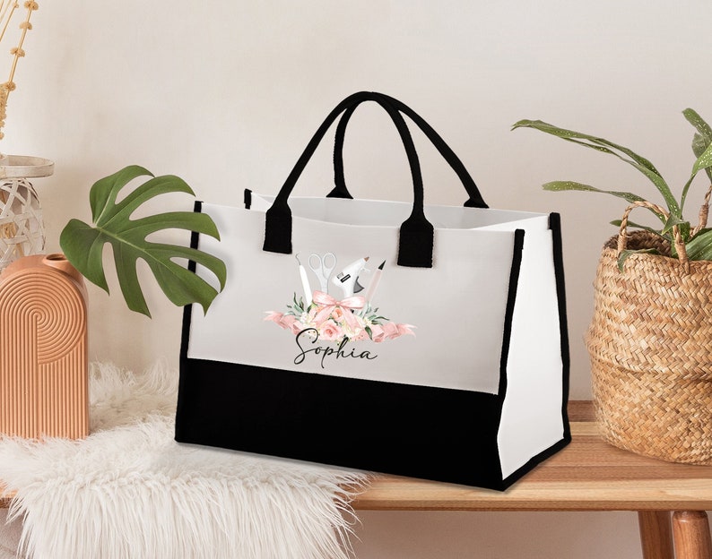 May include: White tote bag with black handles and a black bottom panel. The bag features a floral design with scissors, a glue gun, and the name "Sophia" in script. The bag is sitting on a wooden surface.