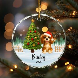 May include: A clear, round Christmas ornament with a festive scene. Inside, a decorated Christmas tree, a Cavalier King Charles Spaniel wearing a Santa hat, and the name "Bailey" with the year "2025". The ornament hangs from a gold string.