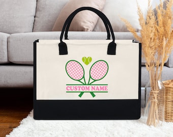Puede incluir: Una bolsa de lona blanca con asas negras y fondo negro. La bolsa tiene un diseño de raqueta de tenis verde con cuerdas rosas y una pelota de tenis verde con un corazón rosa. El texto "CUSTOM NAME" está impreso en negro debajo del diseño.