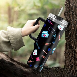 May include: Black stainless steel travel mug with a black handle and a lid. The mug has a pattern of medical symbols, including a stethoscope, bandages, and a nurse's cap. The symbols are in shades of blue, pink, and red.