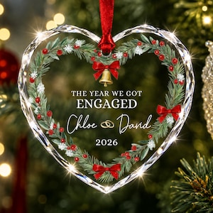 May include: A heart-shaped clear glass ornament with faceted edges, featuring a printed wreath of pine branches, red berries, and red bows. The text reads THE YEAR WE GOT ENGAGED, Chloe and David, and 2026, with a gold bell and red ribbon.