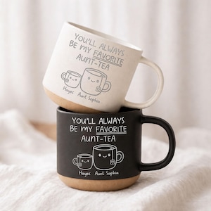 Può includere: Due tazze in ceramica, una color crema e una nera, impilate. Ogni tazza presenta la scritta "YOU'LL ALWAYS BE MY FAVORITE AUNT-TEA" e un simpatico disegno di tazza da tè. Le tazze hanno una base marrone e un manico bianco. È presente anche il testo "Hayes Aunt Sophia".