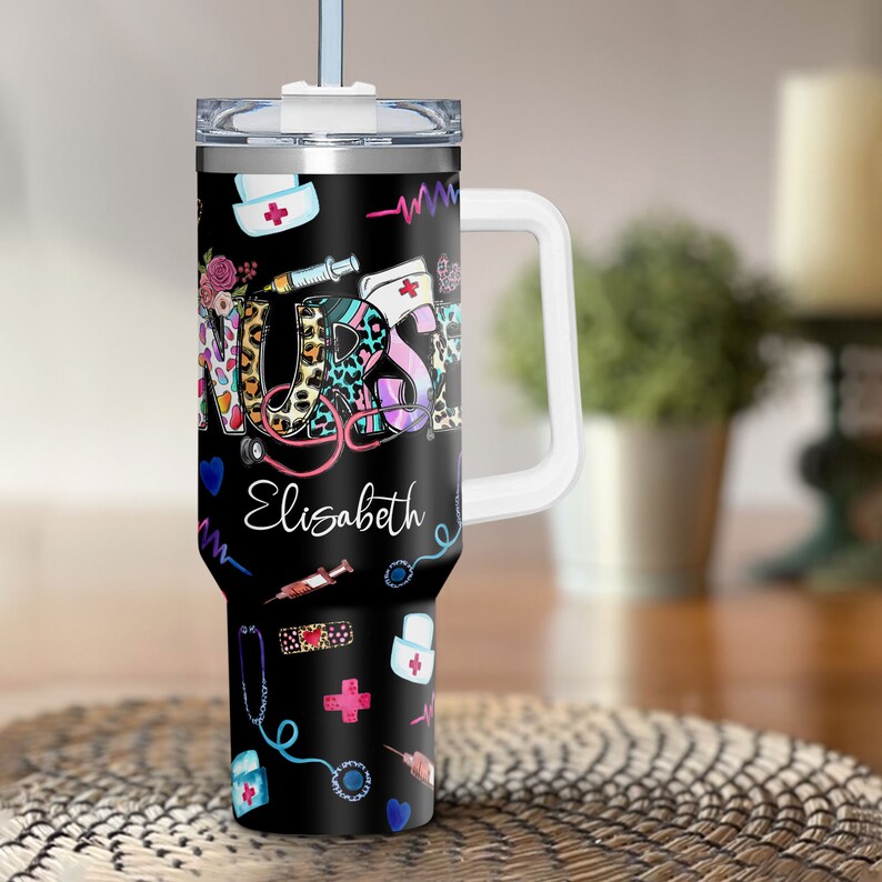 May include: A black stainless steel travel mug with a white lid and handle. The mug features a colorful design with the word "NURSE" and medical symbols, including a stethoscope, a syringe, and a bandage. The name "Elisabeth" is written below the word "NURSE".