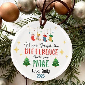 May include: A white ceramic Christmas bauble with a brown leather hanging loop. The bauble displays the text "Never Forget the DIFFERENCE that you MAKE" with festive stockings and Christmas trees. It also includes "Love, Emily 2025".