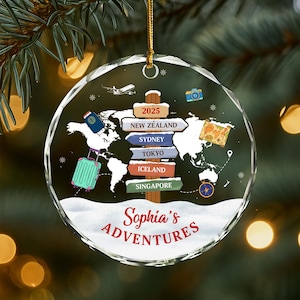 May include: A clear, round Christmas ornament with a world map design. The ornament features a signpost with destinations like New Zealand, Sydney, and Tokyo. A suitcase, passport, and camera are also depicted. The text "Sophia's Adventures" is written in red.