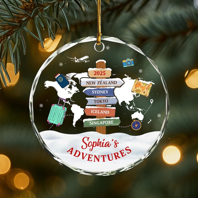 May include: A clear, round Christmas ornament with a world map design. The ornament features a signpost with destinations like New Zealand, Sydney, and Tokyo. A suitcase, passport, and camera are also depicted. The text "Sophia's Adventures" is written in red.