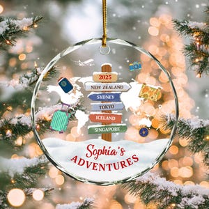 May include: A clear, round Christmas ornament with a gold string. The ornament features a world map design with travel-themed icons and the text "Sophia's Adventures." Destinations include New Zealand, Sydney, Tokyo, Iceland, and Singapore.