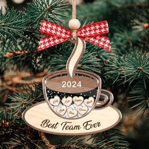 May include: Wooden coffee cup Christmas ornament with the text "2024" and "Best Team Ever." The ornament has a red and white bow and includes names on heart-shaped cutouts. It hangs on a Christmas tree.
