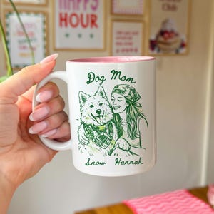 Może przedstawiać: Biały ceramiczny kubek z r&oacute;żowym wnętrzem. Kubek przedstawia zieloną ilustrację kobiety obejmującej psa, z napisem "Dog Mom" powyżej i "Snow Hannah" poniżej. Kubek jest trzymany przez osobę.