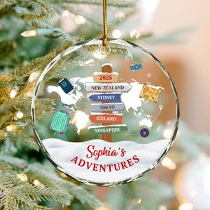 May include: A clear, round Christmas ornament with a gold string. The ornament features a world map design with travel-themed illustrations, including a suitcase, passport, and camera. The text "Sophia's ADVENTURES" is displayed in red, with destination signs pointing to New Zealand, Sydney, Tokyo, Iceland, and Singapore.