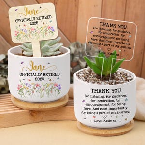 May include: Two white ceramic pots with plants, each on a wooden coaster. One pot has a wooden sign that reads "Jane OFFICIALLY RETIRED 2025". The other pot has a clear sign that says "THANK YOU". Both pots have floral and butterfly designs.